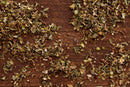 World Spice El Greco by World Spice scattered on a wooden surface, displaying green and brown leafy fragments in various sizes and shapes—an aromatic blend evocative of classic Mediterranean herbs.