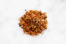 A close-up, top-down view of a small pile of World Spice Fajita & Taco Spice seasoning blend by World Spice, showcasing colorful bits of red chili flakes, dried green herbs, salt, and assorted spices—ideal for infusing bold flavor into ground meat or creating full-flavored fajitas—centered on a pristine white marble surface.