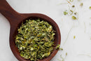 A wooden spoon brimming with World Spice Fenugreek Leaf, an aromatic dried green herb from World Spice, rests elegantly on a white surface, with a few leaves delicately scattered nearby.