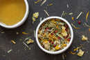 A white bowl filled with World Spice’s Firefly Garden loose herbal tea blend—including dried flowers, green leaves, and red berries—sits on a dark surface next to a cup of brewed tea. Some of the World Spice tea leaves and petals are scattered around.