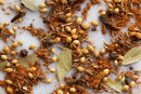 A close-up of World Spice Indian Garam Masala by World Spice shows a vibrant assortment of dried curry spices—coriander seeds, whole black peppercorns, bay leaves, cumin seeds, cardamom pods, cloves, and ground cinnamon—scattered across a white surface.