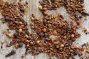 A close-up of a rustic wooden surface scattered with mixed whole and ground aromatic spices, featuring World Spice Kashmiri Garam Masala by World Spice, with visible coriander seeds, peppercorns, roasted masala blends, and other spice fragments.