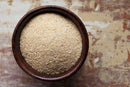 A brown bowl filled with fine, tan-colored breadcrumbs sits on a rustic, textured wooden surface, subtly dusted with World Spice Garlic Granules from World Spice for added flavor.