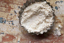 A fluted metal dish brims with flour atop a distressed wooden surface, a light dusting spilling onto the table. Wisps of World Spice Garlic Powder by World Spice are scattered among the flour, hinting at savory creations to come.