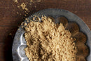 A close-up of World Spice Ginger Powder, finely ground and displayed on a decorative metal plate with some powder scattered onto a wooden surface—perfect for adding warmth and depth to marinades or baking recipes.