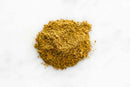 A small pile of World Spice Jerk Seasoning—a finely ground, yellowish-brown blend from World Spice, perfect for authentic Caribbean barbecue jerk—sits on a white surface.