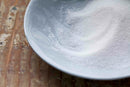 A close-up of World Spice Citric Acid, the signature tangy crystals from World Spice, sprinkled over white granulated sugar on a light blue plate resting on a wooden surface.