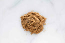 A small pile of World Spice Chaat Masala by World Spice rests on a white marble surface, displaying a light brown color with a slightly uneven texture and a few scattered granules around the edges.