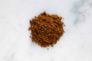 A small mound of finely ground, dark brown aromatic World Spice Kala Masala—a traditional Maharashtrian spice blend from World Spice—sits on a white marble surface.