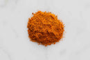 A small pile of World Spice Tikka Masala, a bright orange-brown powdered spice blend from World Spice, rests on a white surface—evoking the vibrant seasonings classic to British dishes like Tikka Masala.