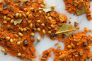 A close-up of World Spice Tikka Masala by World Spice, featuring a vibrant orange-red spice blend accompanied by whole coriander seeds and bay leaves, reminiscent of classic British cuisine favorites like Tikka Masala, scattered on a light surface.