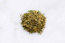 A small pile of World Spice Orange Tarragon, a French seasoning blend from World Spice, shows off mixed dried herbs and spices with green, yellow, brown, and subtle orange flecks of tarragon on a white marble surface. This blend delivers a delicate floral flavor in every sprinkle.