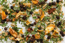 A close-up of World Spice Orange Tarragon by World Spice reveals a vibrant dried herb and spice blend, reminiscent of classic French seasonings, featuring green leaves, black peppercorns, orange peel pieces, and dried onion or garlic atop a white surface. The aromatic medley promises a subtle floral flavor with hints of tarragon and orange throughout.