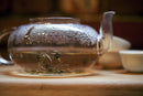 A clear glass teapot filled with World Spice Jasmine Pearl tea leaves brews aromatic flavors on a wooden surface, with condensation visible inside the teapot. In the background, white teacups from World Spice are softly blurred.