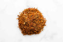 A small mound of World Spice Ras El Hanout by World Spice rests on a white surface, showcasing a blend of red, brown, and orange hues—this savory spice mix captures the artistry found in a traditional spice shop.