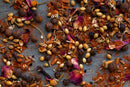 A close-up of assorted spices—including coriander seeds, cloves, peppercorns, rose petals, and ground red spice—scattered across a grey surface, capturing the vibrant essence of World Spice Ras El Hanout by World Spice.