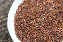 A close-up of World Spice Rooibos loose tea leaves from World Spice, known for their rich antioxidants and naturally caffeine-free qualities, showcases their reddish-brown color on a white plate with a wooden surface in the background.