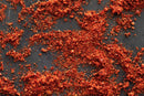 World Spice Rooster Spice by World Spice, with its vibrant red chili powder and flakes, is scattered across a dark surface, creating a textured, fiery effect reminiscent of Southeast Asian cuisine ingredients, with uneven piles and crumbs spread out randomly.