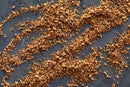 Scattered across a dark gray surface, loose, golden-brown crumbs of World Spice Sassy Steak Spice by World Spice form an uneven, diagonal pattern. Both fine and coarse textures are visible, and the blend is infused with a hint of Vietnamese cinnamon.