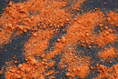 World Spice America's Seasoned Salt by World Spice, in vibrant orange-red powder form, is scattered across a dark surface, with visible small clumps and granules of varying sizes.