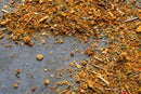 Close-up of scattered World Spice Thai Seasoning by World Spice on a gray surface, displaying an array of colorful flakes, seeds, and small pieces—featuring hints of ginger and classic Asian-inspired herbs and spices often used in marinades.