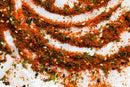 World Spice Togarashi by World Spice—an aromatic blend featuring red chili powder, sesame seeds, and dried green herbs—scattered in swirling patterns on a white surface, reminiscent of the vibrant Japanese condiment Togarashi and perfect for sprinkling over Japanese noodles.