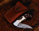The Texan Knives Personalized Damascus Steel Pocket Knife features a patterned blade and wood-and-bone handle, paired with a brown leather sheath—making it the perfect custom Valentine’s Day gift knife for your husband.