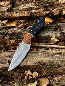 The Texan Knives Handmade 8" Damascus Skinning Knife with a buffalo horn and rosewood handle rests on weathered logs scattered with dry leaves, its patterned high-carbon steel blade gleaming beside its leather sheath.