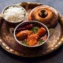 A brass tray holds a bowl of chicken curry, infused with The Spice Lab Organic Ground Turmeric and garnished with cilantro and a dried chili, accompanied by a bowl of white rice and topped with a brass lid, all arranged on a dark surface.