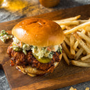 A fried chicken sandwich with pickles and slaw on a bun sits on a wooden board next to a serving of seasoned French fries, each bite bursting with Caribbean-inspired flavor thanks to The Spice Lab Scotch Bonnet Sea Salt by The Spice Lab.
