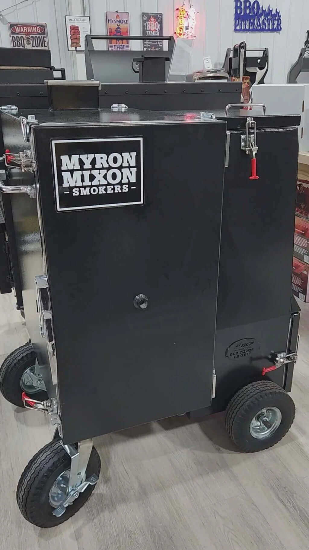 A large black Myron Mixon smoker on wheels sits indoors on a light wood floor. The smoker has a front door with a latch, a Myron Mixon Smokers logo, and robust handles for transport. BBQ-themed signs decorate the background.