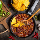 A hearty pan of chili made with kidney beans and ground meat, expertly seasoned with The Spice Lab Organic Chili Powder, is garnished with crispy tortilla chips. On the rustic wooden table beside it are extra tortilla chips, sliced jalapeños, whole chili peppers, garlic cloves, and a fresh lime for added flavor—courtesy of The Spice Lab.