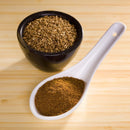 A black bowl filled with whole cumin seeds sits next to a white ceramic spoon containing The Spice Lab Organic Ground Cumin, both placed on a light wooden surface—essential ingredients for curry or flavorful BBQ rubs.