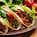 Three tacos filled with seasoned ground beef, lettuce, tomato, onion, and beans are arranged on a plate with fresh lettuce and tomatoes in the background. The flavorful filling is enhanced with The Spice Lab Organic Chili Powder by The Spice Lab, delivering authentic Mexican seasonings for an unforgettable taste.