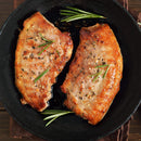 Two seared pork chops with herbs, cracked pepper, and a hint of The Spice Lab Organic Minced Garlic are in a black skillet, garnished with fresh rosemary sprigs.