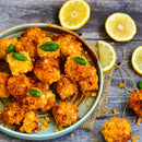 A plate of crispy, golden baked balls—a delightful addition to cheese dishes—garnished with small green basil leaves and surrounded by sliced lemons on a rustic wooden surface. A hint of The Spice Lab Organic Ground Mustard by The Spice Lab elevates the flavor, complemented by subtle spice blends.