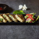 Four pieces of stuffed, grilled quesadilla are arranged on a black tray beside fresh vegetable slices, including tomatoes, cucumbers, and green chili, with a garnish of The Spice Lab Whole Leaf Cilantro by The Spice Lab on a gray stone surface.