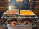 Two Pimp My Grill® Gen2 Slide-Out Rack System racks inside a pellet grill, holding aluminum pans of baked beans and macaroni and cheese on top, and whole roasted chickens and corn on the cob on the bottom.