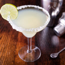 A margarita in a glass with a salted rim of The Spice Lab Traditional Margarita Salt Cocktail Rimmer and a lime wheel garnish sits on a wooden table next to a cocktail shaker, spoon, and cocktail rimmer from The Spice Lab.