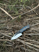 The Texan Knives Hunters Knife features a 9" D2 steel blade and an epoxy pine cone resin handle. Shown outdoors on dry twigs and grass, this handcrafted knife includes a durable leather sheath.