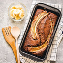 A loaf of chocolate banana bread in a pan, topped with banana slices and a sprinkle of The Spice Lab Organic Ground Allspice, sits on a checked towel alongside a bowl of sliced bananas and wooden kitchen utensils.