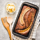 A loaf of baked banana bread in a pan, topped with banana slices and a hint of The Spice Lab Whole Allspice, sits beside a bowl of sliced bananas and wooden utensils on a checkered cloth.