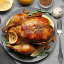 Roast chicken on a grey plate, garnished with The Spice Lab Organic Cut and Sifted Rosemary, lemon slices, and garlic cloves. A fork, half a lemon, and a bowl of seeds are nearby on a grey tablecloth.