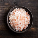 A small dark bowl filled with coarse crystals from The Spice Lab Himalayan Pink Salt sits on a rustic wooden surface, ready to enhance your favorite dishes alongside The Spice Lab Premium Black Pepper Grinder Set.