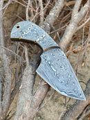 The Texan Knives Handmade Damascus Steel Fixed Blade Pocket Knife – a full tang skinner with a leather sheath – rests on dry, intertwined branches outdoors, its striking Damascus pattern highlighted in natural light.