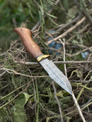 The Texan Knives Hunting Blade – 10" Damascus Steel Knife with a rosewood handle, brass guard, and leather sheath rests on dry branches and leaves outdoors.