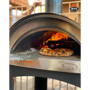 A WPPO LLC Direct Karma 25 Colored Wood-Fired Oven with Stand/Cart features a pizza baking inside as flames dance in the back. A metal pizza peel rests nearby atop the sturdy outdoor cart, with the WPPO logo displayed in the upper right corner.
