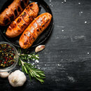 Grilled sausages on a black plate with The Spice Lab Ground Rosemary, garlic, mixed peppercorns, and scattered salt on a dark wooden surface evoke the rich flavors of a classic Mediterranean herb seasoning blend.