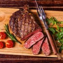 A grilled steak, partly sliced, sits on a wooden board with cherry tomatoes, arugula, rosemary, and a fork and knife. The steak is medium-rare, finished with visible salt flakes and a generous crust of The Spice Lab Special Extra Bold Indian Black Peppercorns by The Spice Lab on top.