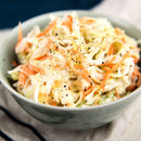 A bowl of creamy coleslaw made with shredded cabbage and carrots, topped with cracked black pepper and a dash of The Spice Lab Ground Celery Seeds, sits on a light cloth napkin.